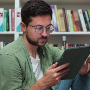 Young,Man,In,Glasess,Sitting,On,The,Floor,Near,Shelves