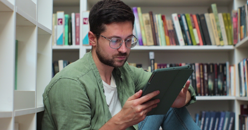 Young,Man,In,Glasess,Sitting,On,The,Floor,Near,Shelves